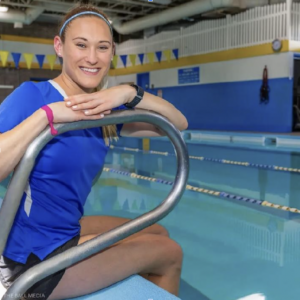 Photo of Janine, swimming instructor at Worcester Fitness. She is crouched next to the railing of an indoor pool. She is wearing a blue short sleeve shirt and black shorts